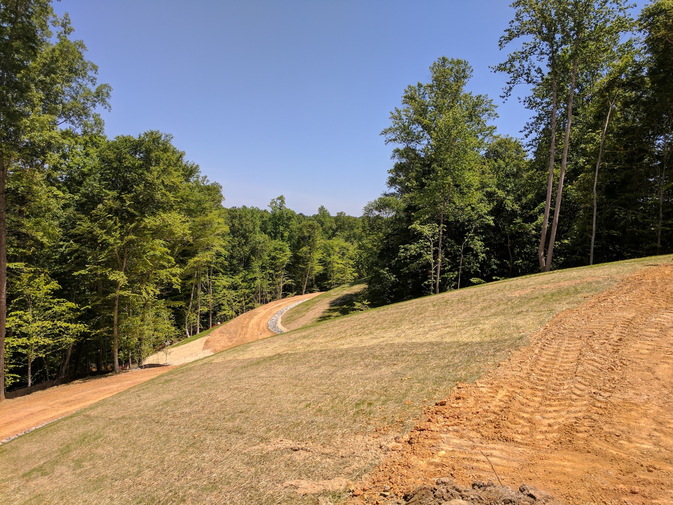 Land development site with graded earth, grass, and tree-lined access road under a clear sky